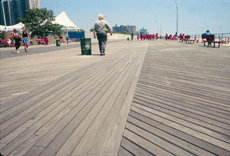 Boardwalk in foreground and amusements in the background and people walking, including a Parks Department employee in khaki and green, on the boardwalk.