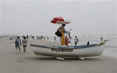 A small boat on a beach, the word "Wildwood" on its side, with a lifeguard on a stand next to the boat and people walking on the beach